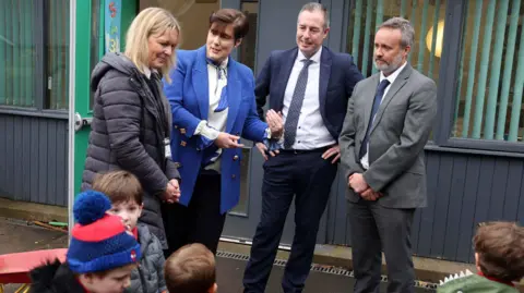 Department of Education Paul Givan, wearing a dark blue suit, white shirt and dark tie stands with his hands on his hips outside a school. Standing beside him is Irish Education Minister Norma Foley wearing a bright blue jacket and white and blue blouse. She is talking and gesturing with her hands. They are talking to a woman with blonde hair and a grey-haired man in a suit. Four children are in the foreground, partially out of view.