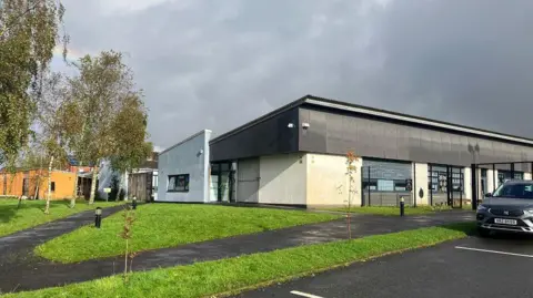 Exterior view of Bunscoil an tSléibhe Dhuibh in west Belfast. It is a modern building surrounded by some trees, a lawn and paved walkways. A car is parked in its carpark.