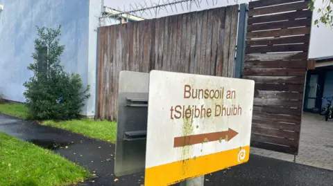 A white, yellow and brown sign pointing to the entrance of Bunscoil an tSléibhe Dhuibh. It is located in front of a wooden barrier with anti-climb security spikes on the top of the wood.