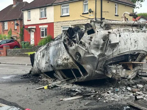 PA An upturned burnt out car in front of houses.