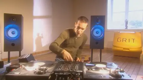 Martyn Bennett sits at a vinyl mixing deck, flanked by large speakers on stand in a room with a large yellow street grit bin in below the window. a statuette is casting a shadow of a waving figure on the wall.