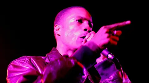 Getty Images D Double E holds a microphone to his lips as he performs on stage. He is wearing a leather jacket and looking away to the right of the camera towards the audience.