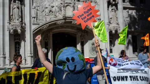 Carl Court/Getty Images A woman with a large Planet Earth mask obscuring her face celebrates while holding a sign saying "The fight for our future" outside the High Court in London. Protesters in the background also hold signs and banners