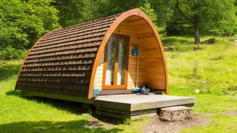 Getty Images A wooden hut with a point roof sits in a field. There are glass double doors on the front and a number of blue brushed hanging up. In the background you can see bright green grass and trees.
