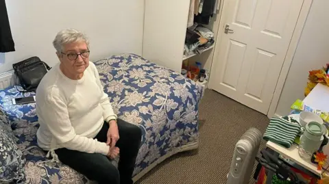 Emma Curtis Susan Curtis - a woman in her 70s with short grey hair - sitting on a bed in a small room