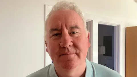 BBC A close up photo of Pilib Mistéil, principal of Bunscoil an tSléibhe Dhuibh in Belfast, looking to camera wearing a light-green/grey top
