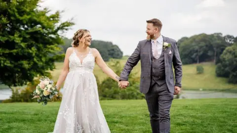 Charlotte and Dave Lay A newlywed couple hold hands and smile at each other against a backdrop of hills and trees. The groom is wearing a grey suit and the bride, who is holding some flowers in her other hand, is wearing a traditional white dress.