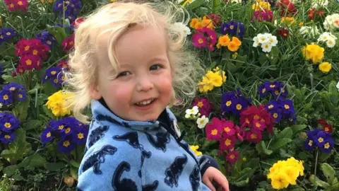 Police Scotland Young boy with shoulder length blonde hair, wearing a blue fleece sweater with dinosaurs, smiling standing in front of a patch of flowers.