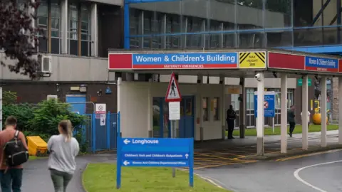 PA Entrance of hospital - sign reads Women & Children's Building - a man with a rucksack and a woman in a grey sweater are walking towards the door