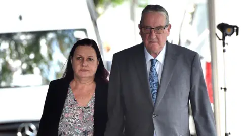 PA Media Susan Letby, with dyed red hair and wearing a black blazer over a flowery blouse, and John Letby, who has grey hair, glasses and wears a grey suit with a blue tie, walk towards the camera with a serious expression