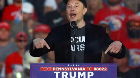 Reuters Elon Musk, wearing a black 'Make America Great Again' cap, a black blazer and grey emblazoned t-shirt, points to a sign for Donald Trump's presidential campaign as he speaks at a Trump rally in Pennsylvania