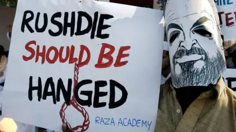 Getty Images An Indian Muslim wears a mask of Indian writer Salman Rushdie as he displays a placard condemning Rushdie during a protest in Bombay, 12 January 2004. During the protest against Rushdie's presence in the city, organised by several Muslim organisations, a reward of Rs 100 000 (2 199 USD) to anyone who blackens the face of Rushdie,who in his book The Satanic Verses allegedly made remarks against Islam's holy prophet Mohammed. (Photo by SEBASTIAN D'SOUZA / AFP) (Photo by SEBASTIAN D'SOUZA/AFP via Getty Images)