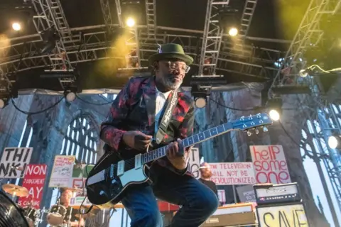 Getty Images Lynval Golding seen wearing a hat and red jacket playing a guitar during a gig in 2019 with The Specials in the ruins of Coventry Cathedral.