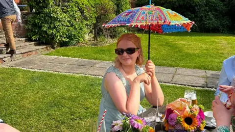 Handout Isobel, pictured sitting outside at a wooden picnic table, holding a colourful parasol above her head. She is wearing a light blue summer dress and sunglasses. A bunch of flowers and some drinks can be seen on the table in front of her.