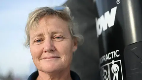 Getty Images Pip Hare poses for a photograph aboard her Imoca monohull "Medallia" at the starting site of the 10th edition of the Vendee Globe around the world monohull solo sailing race, at the Vendee Globe village in Les Sables-d'Olonne, western France. Her blonde/grey hair is tied back and she is wearing a navy jumper.
