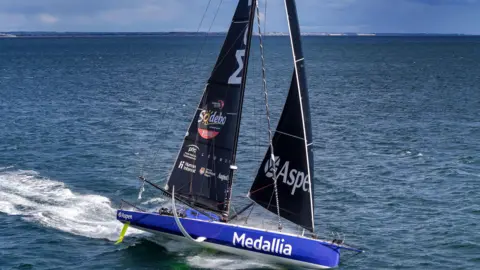 PA Media A drone image of the Medallia yacht sailing on the sea. It has large black sails and a blue body. The name Medallia is printed on the side of the yacht's body in white lettering.