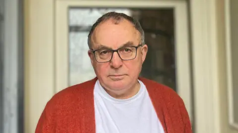 Andrew in a white T-shirt and orange cardigan standing on his doorstep looking into the camera