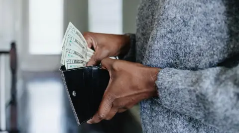 Getty Images A cropped image showing the hands of a woman holding a wallet and fanning out $20 bills
