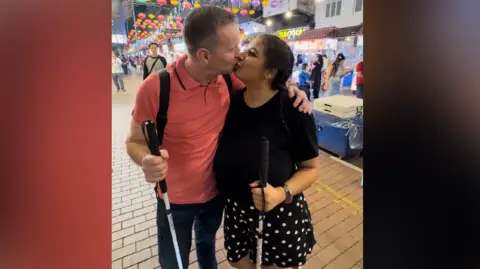 Nicola Thomas A man and woman kissing on a street in east Asia. They are both holding canes as they have visual impairments. The man is wearing blue jeans and a salmon pink shirt, the woman is wearing a black top and a black and white polka dot skirt.