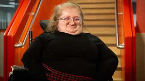 A woman in a wheelchair, smiling at the camera. She is wearing a black top with a black and red skirt in a houndstooth pattern. There are stairs behind her with the banisters painted orange.