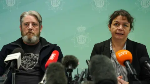 PA Media A sombre looking Tim Edwards, with grey hair and a grey beard, sitting in front of a series of microphones alongside Det Supt Sue Coombs, who has black hair tied into a bun and wears a black blazer