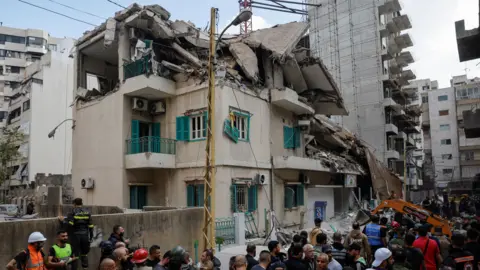 Reuters The roof of a multi-story building with tan walls and green balcony railings and window shutters is collapsed from an air strike, with a crane and a crowd of rescuers and onlookers below