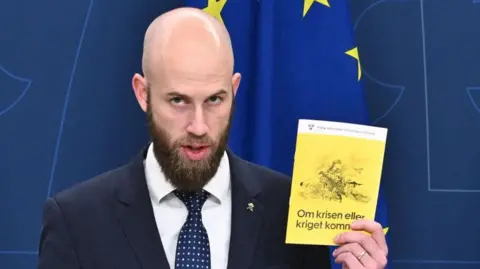 Getty Images Sweden's Minister for Civil Defence Carl-Oskar Bohlin holds a copy of the new version of the preparedness booklet "If the crisis or war comes". The pamphlet has a yellow cover and an image of a woman in army fatigues holding a large gun.
