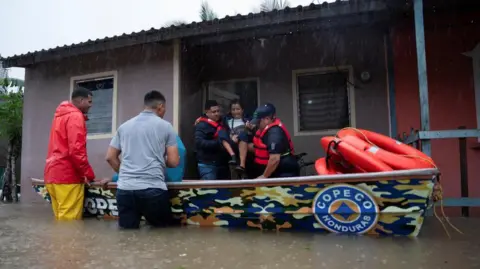 Reuters Rescue workers wearing red inflatable jackets carry a woman aloft into a waiting boat. Rain is falling and the road is flooded with water reaching the rescuers' thighs.