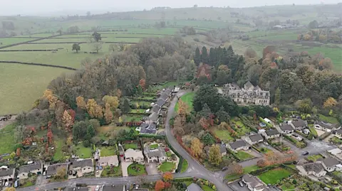The picture is an aerial image high above the town of Bakewell in the Derbyshire Dales. It shows a number of homes with trees and green fields stretching out into the distance.