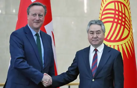 Getty Images Mr Cameron (L) and Mr Kulubaev (R) shake hands while standing in front of the British and Kyrgyzstan flags.