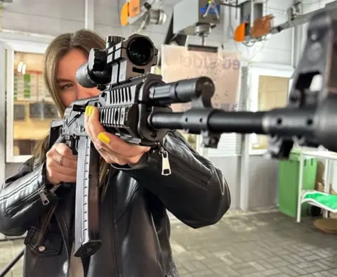 Instagram A young woman with long brown hair poses with an automatic rifle in what could be a shooting range. She is looking through the telescopic lens, with the muzzle of the gun facing the camera. She has bright yellow nail varnish and wears a leather jacket.