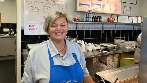 Sharon Foy Sharon Foy owner of Sharon's Plaice chip shop stood behind the counter looking at the camera wearing a blue pinafore