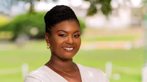Tremaine Yearwood Brittney McKenzie, wearing gold hoop earrings, a gold necklace and a white cardigan, poses for a photo