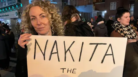 Donna McCarthy has long curly blond hair and is holding a sign saying Nakita the warrior while other women at the protest stand behind her.