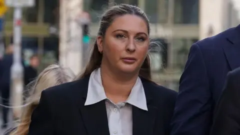 PA Media Ms Hand has long blonde hair and is wearing makeup. She is wearing a black suit jacket with a blue shirt.