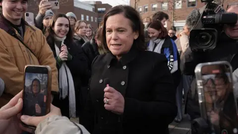 PA Media Mary Lou McDonald wearing a black button-up coat at an election event in Dublin. Two people are taking pictures of her with their phones while a cameraman is behind her.