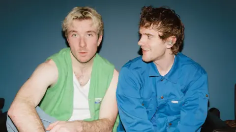 Lauren Harris Good Neighbours made up of of (L-R) Oli Fox and Scott Verrill. Oli wears a green vest-cardigan over a white vest top, his hands in his lap as he sits cross-legged. Scott sits beside his bandmate and looks across at him, smiling, while wearing a blue overshirt.