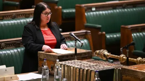 PA Media Rachel Hopkins wearing a black jacket over a red dress, asking a question in the House of Commons, she has dark long hair, glasses and has a necklace on. The seats are green leather, she is talking into a microphone and is holding a sheet of paper.