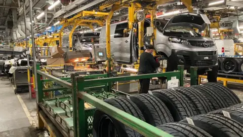Sam Read/BBC A worker in a Vauxhall Luton plant, working on a van, with tyres and car parts around him. There is a lot of machinery, hanging from the ceiling and more vans in a production line.