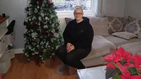 Sidony West Sidony West, a blonde woman with glasses, sits in her living room on a sofa next to a Christmas tree.