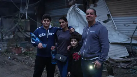 Car dealer Mohamad Marouf (R), younger son, wife and older son (L) standing in front of the destruction on their street from Israeli air strikes. Mr Marouf is holding his mobile phone with the torch on - both sons also have phones in their hands. Older son has one arm wrapped around a water bottle, and the other around his mother's shoulders. She is also holding a water bottle.