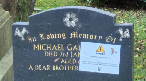 BBC A black gravestone with gold lettering at Riddrie Park Cemetery - the first name of the deceased is Michael, but the surname has been covered by a large sticker saying WARNING with a exclamation mark in a yellow box