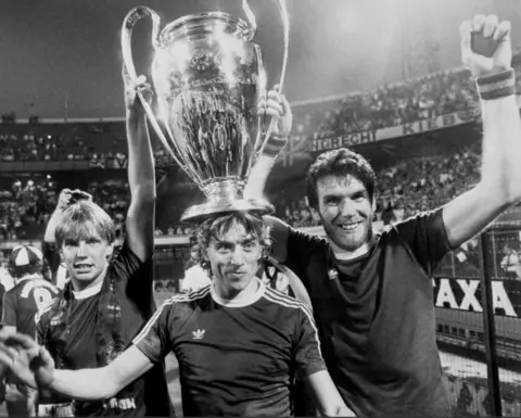 PA Media A black and white photo of three smiling footballers, in football shirts, holding a giant trophy aloft. Football crowds can be seen in the background