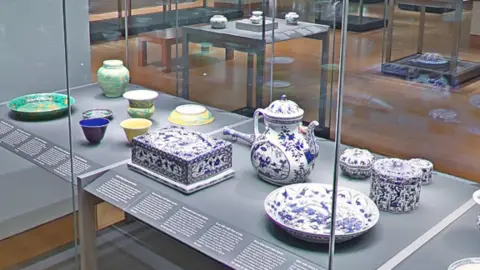 Google Bowls, a jug, and ceramic boxes in white and blue and green and yellow sit in a display cabinet in the British Museum's Chinese ceramics collection