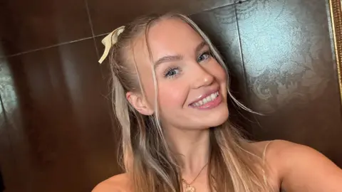 Sophie Jones Sophie is taking a selfie. She has long blonde hair with half of it tied back with a yellow bow holding it in place. She wears a gold necklace and a yellow strappy top.