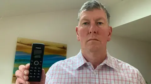 Supplied Paul Toplass stands in his office holding a black portable telephone handset. He has short grey hair and wears a white and pink checkered shirt.