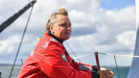PA Media Pip Hare looks over the edge of her yacht toward the sea as she fastens a rope around a metal pole. Her blonde and grey hair is windswept and she is wearing a large red waterproof coat.
