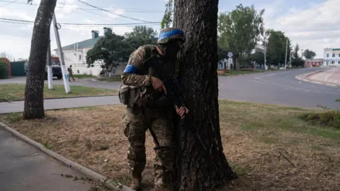 Getty Images A Ukrainian soldier fighting in Russia's Kursk border region. He is carrying an assault rifle and is hiding behind a tree, while wearing a helmet and military fatigues.