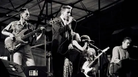 Getty Images The band the Specials play at a concert at Coventry's Butts Stadium. Bass player Horace Panter is at the far left of the picture next to lead singer Terry Hall who is singing into a microphone. Rhoda Dakar sings into a microphone next to Lynval Golding and Jerry Dammers of the band.
