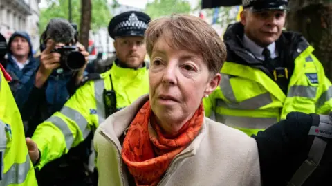 Getty Images Former Post Office CEO Paula Vennells, wearing an orange scarf and beige coat and flanked by police officers in yellow high-vis coats, arrives to give evidence at the Horizon IT public inquiry on Wednesday, 22 May 2024.